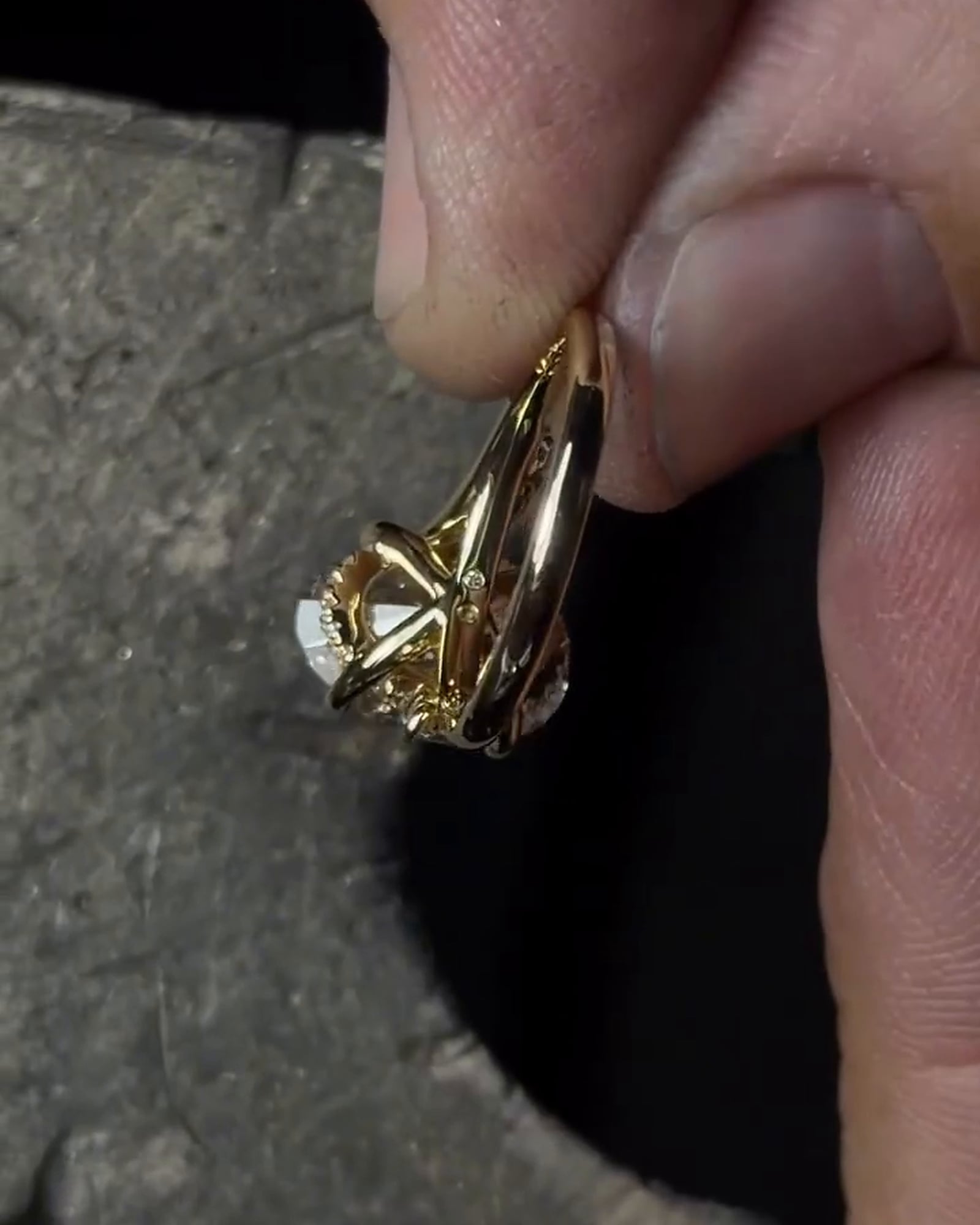 Side profile of a gold-tone ring held between fingers, showing its raised setting, open gallery and curved band.