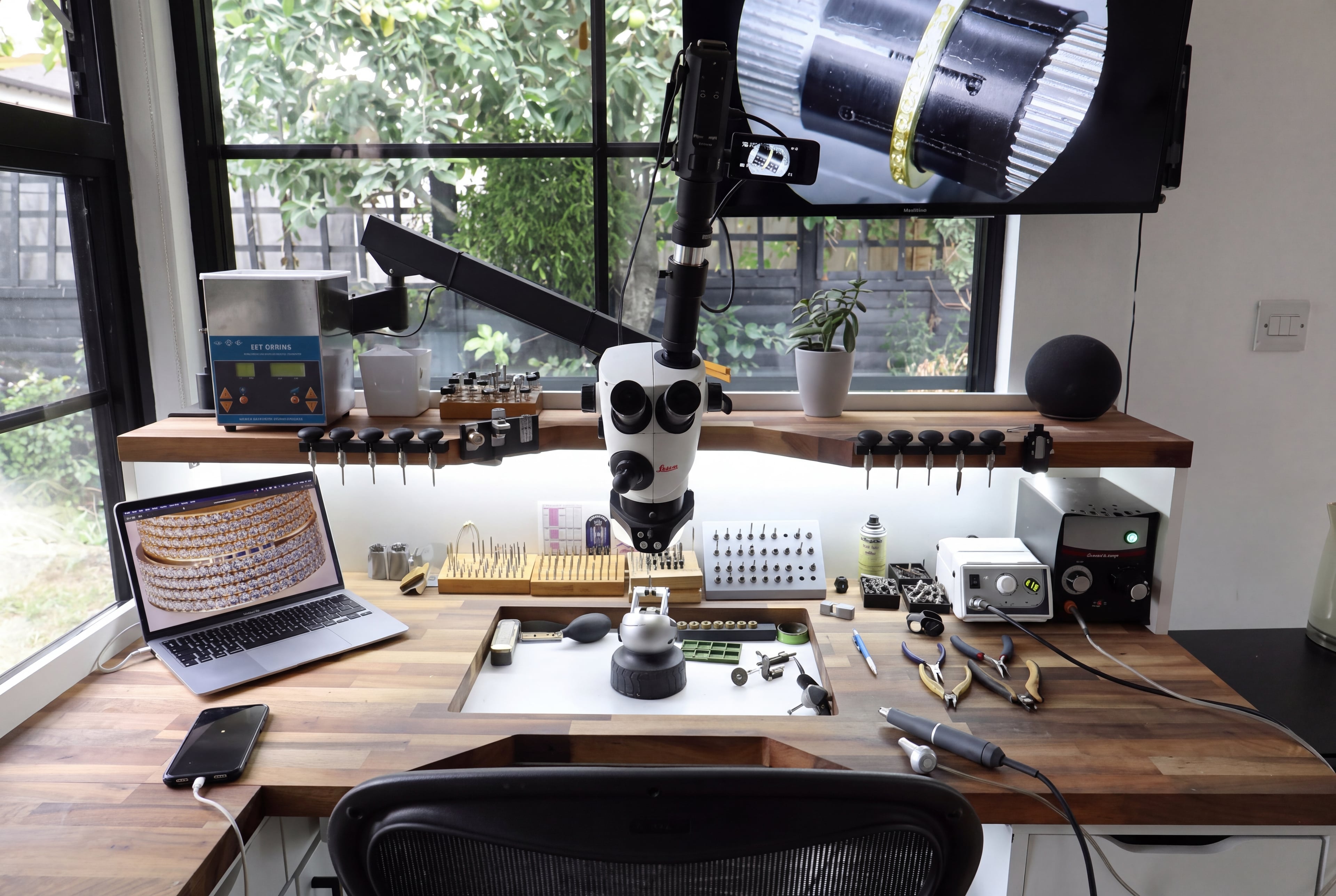 Jewelry workbench with a microscope, organized hand tools and a laptop beside a window.