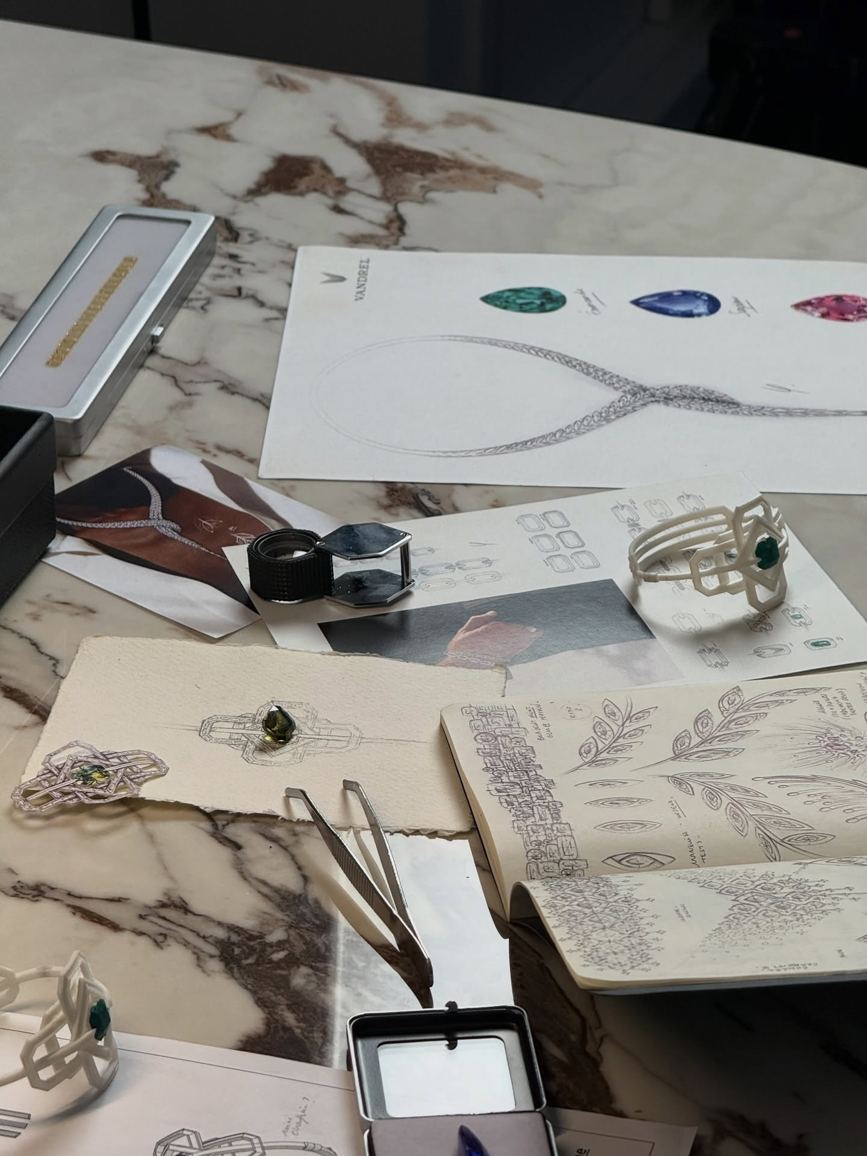Jewelry sketches, pale ring models, colored stones, tweezers and a loupe arranged on a marble table.