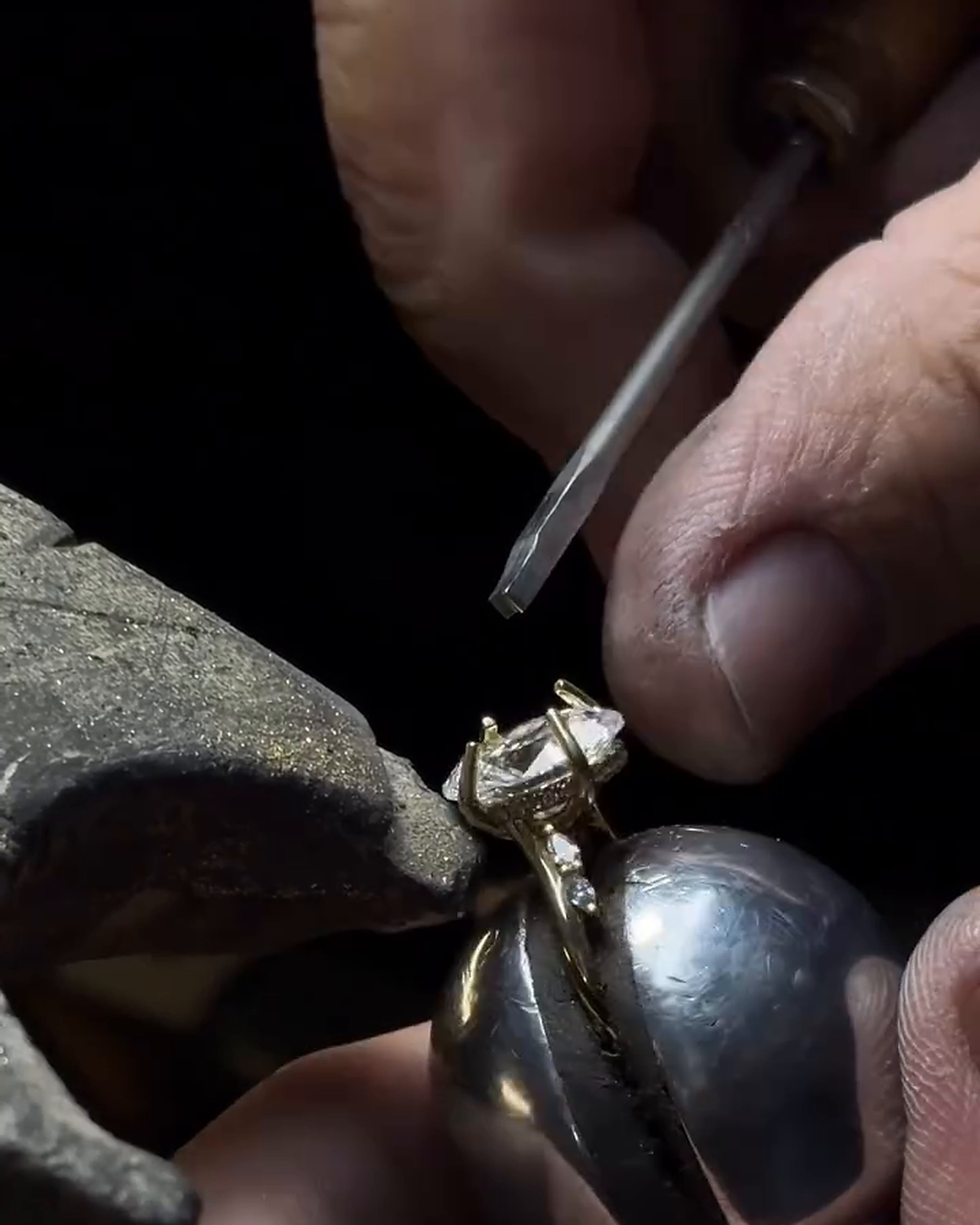 Fingers and a pointed hand tool beside a clear stone in a gold-tone ring setting secured in a bench holder.