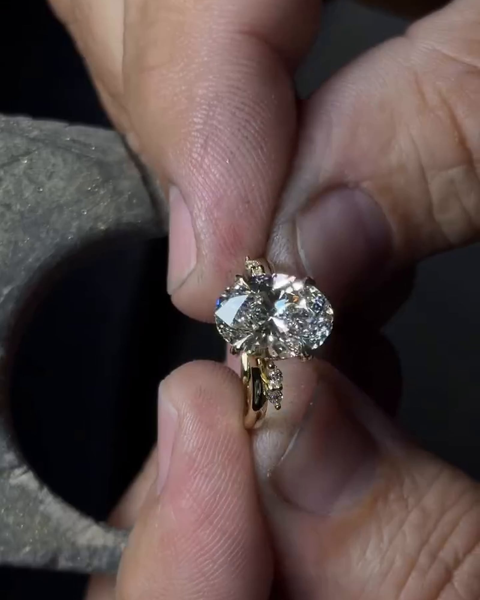 A gold-tone ring held between fingertips, showing a clear oval stone, surrounding prongs and smaller shoulder stones.
