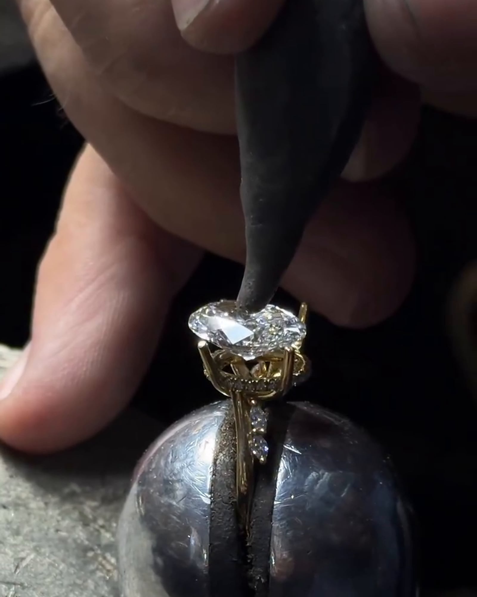 A hand tool touches a clear faceted stone seated in a gold-tone ring setting supported by a metal holder.