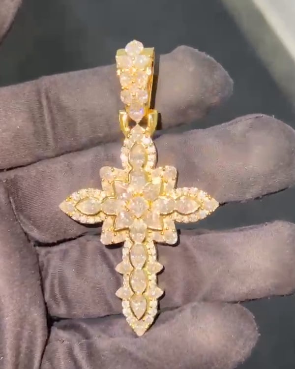 Decorative gold-tone cross pendant with pointed oval stone shapes and a tapered bail resting on a gloved hand.