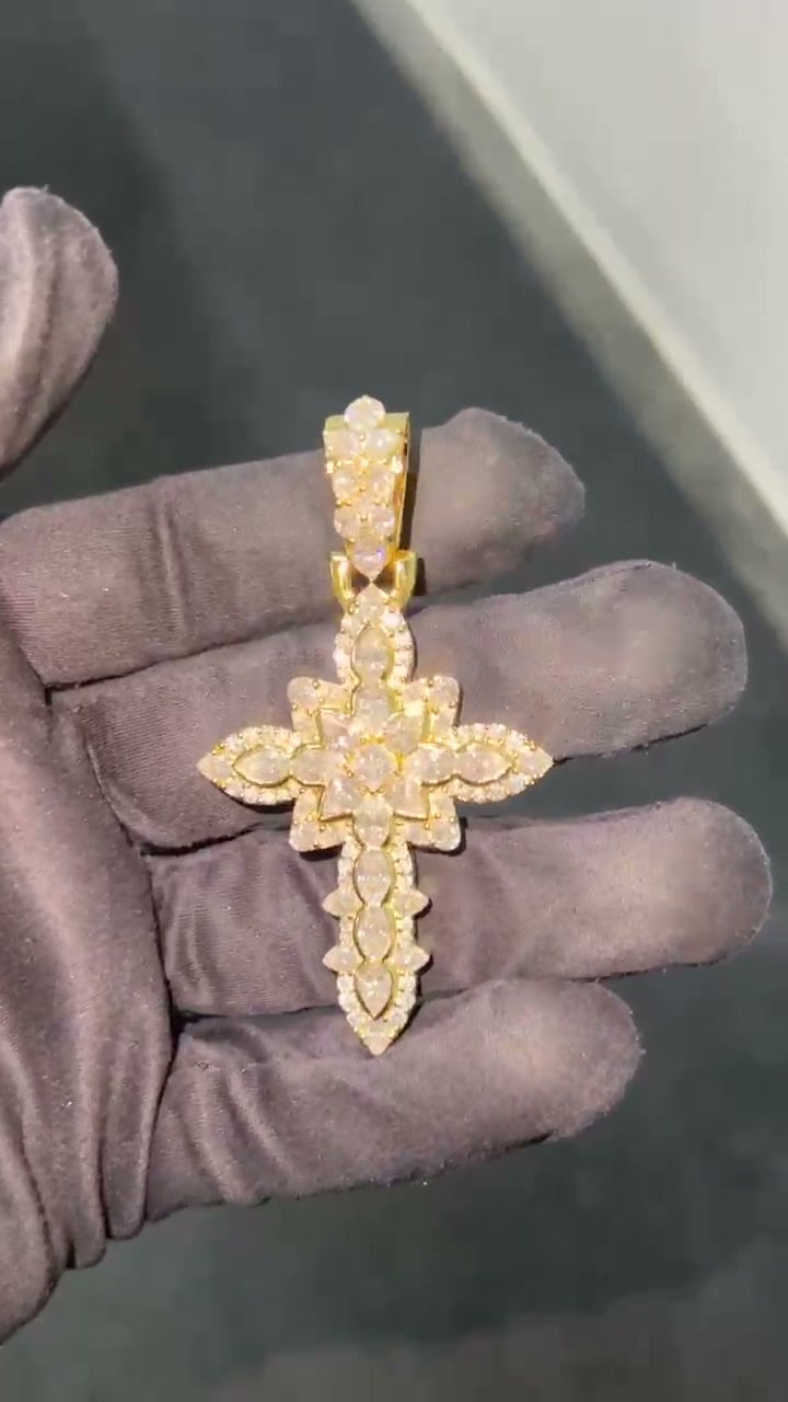 Decorative gold-tone cross pendant with pointed oval stone shapes and a tapered bail resting on a gloved hand.