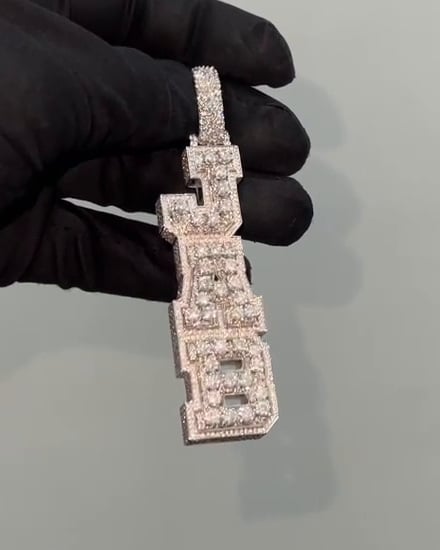 Elongated stone-set pendant formed from stacked block lettering and a broad bail, held by a black glove.