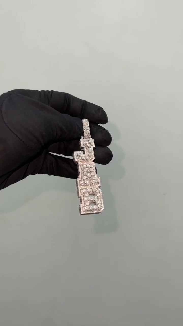 Elongated stone-set pendant formed from stacked block lettering and a broad bail, held by a black glove.