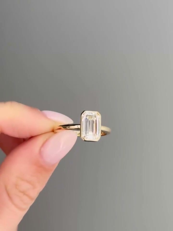 Rectangular step-cut ring enclosed by a yellow-tone bezel, held against a gray background.