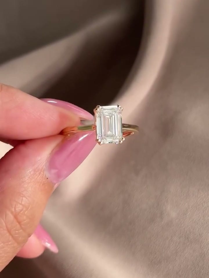 Rectangular step-cut solitaire ring with corner prongs and a slender yellow-tone band, held over fabric.