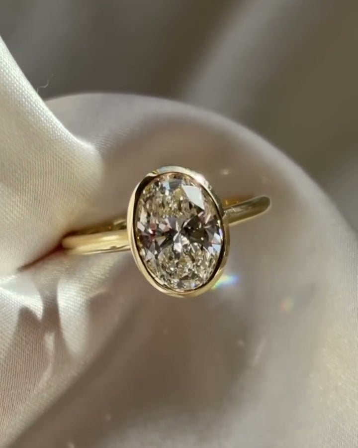 Oval ring with a continuous yellow-tone bezel and slender band on pale satin.