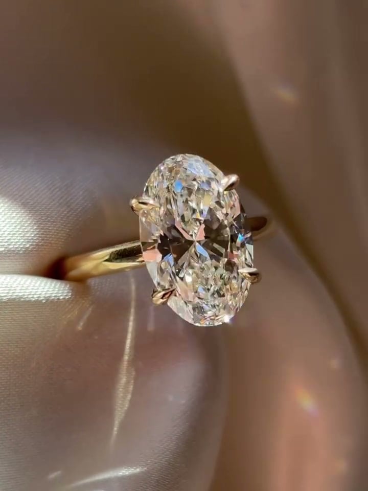 Oval solitaire ring with visible prongs and a slender yellow-tone band resting on pale satin.