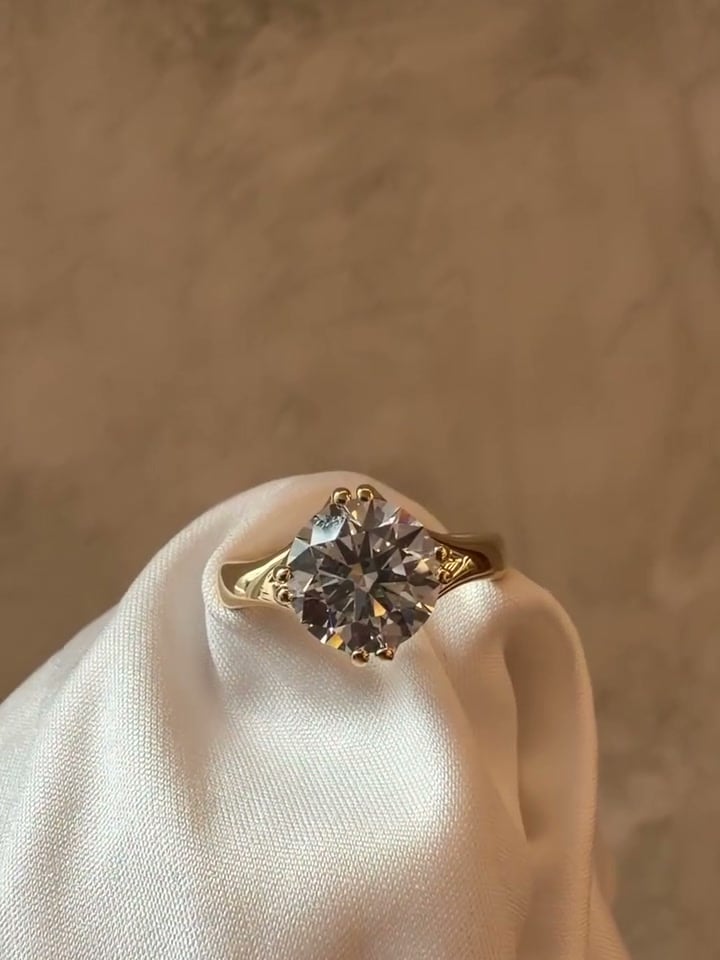 Round solitaire ring in a yellow-tone prong setting with curved shoulders, supported by pale satin.