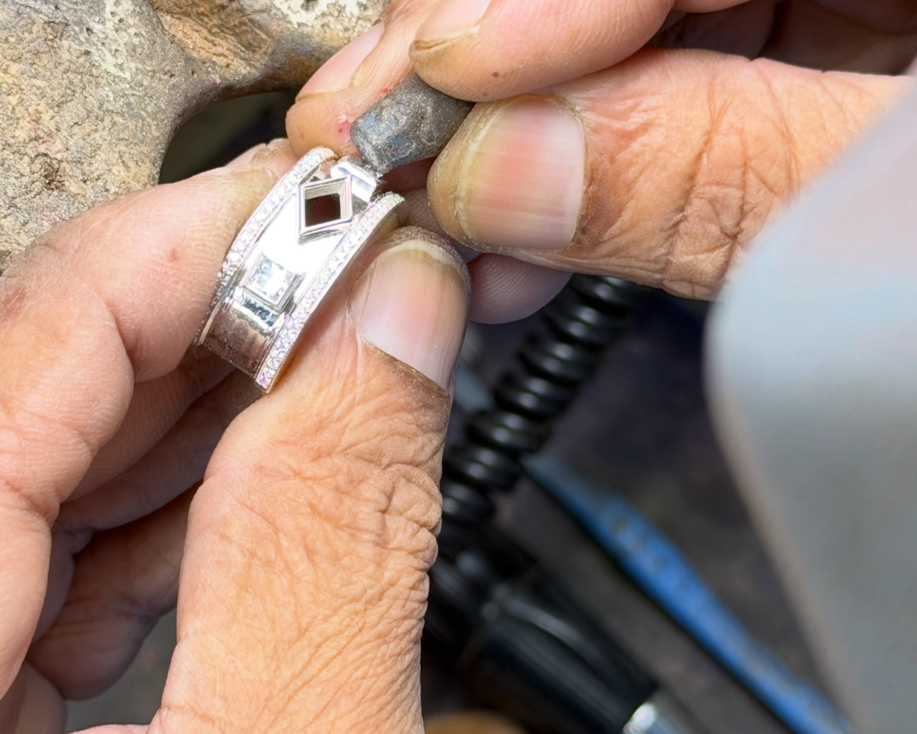 Fingers hold a silver-tone jewelry mount while a small hand tool meets its edge.