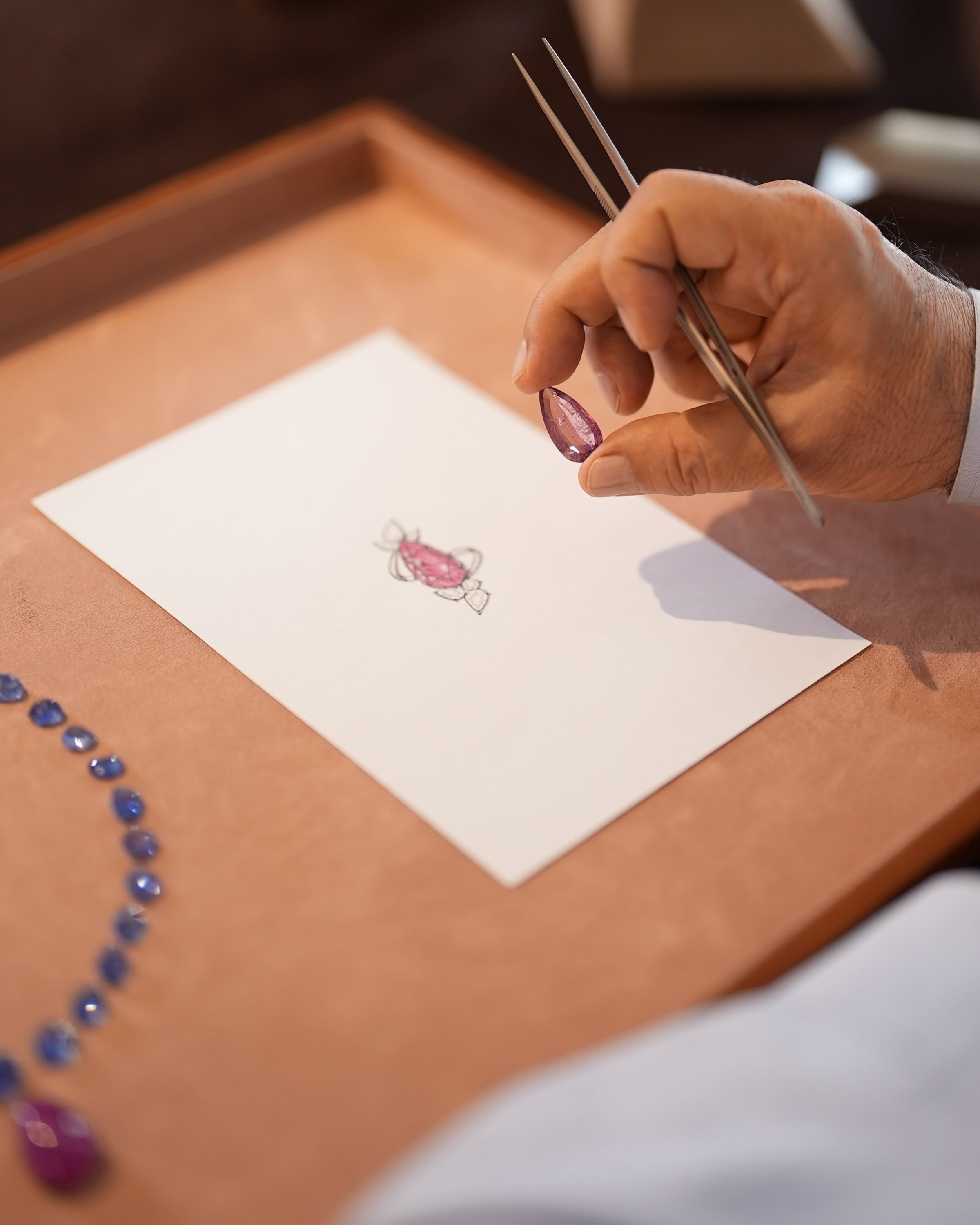A hand holds a fine brush above a colored jewelry design, with loose stones beside the paper.